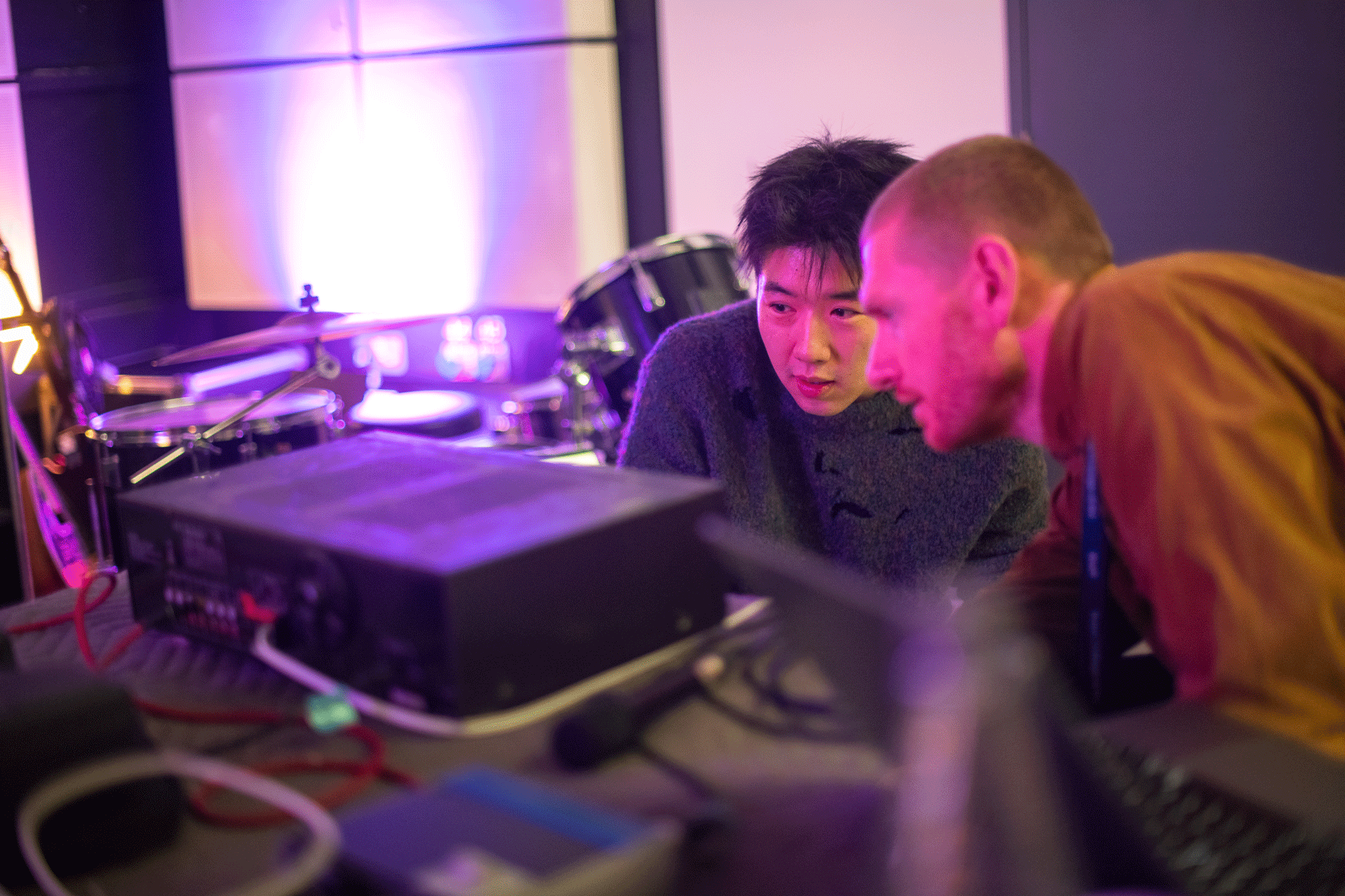Two students in the sound lab examining technology with a drum kit in the background