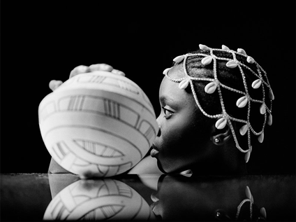 A black and white photo of the photographer's head in profile, wearing a headress made of sheels, with a ceramic vessel used in  Yoruba culture.