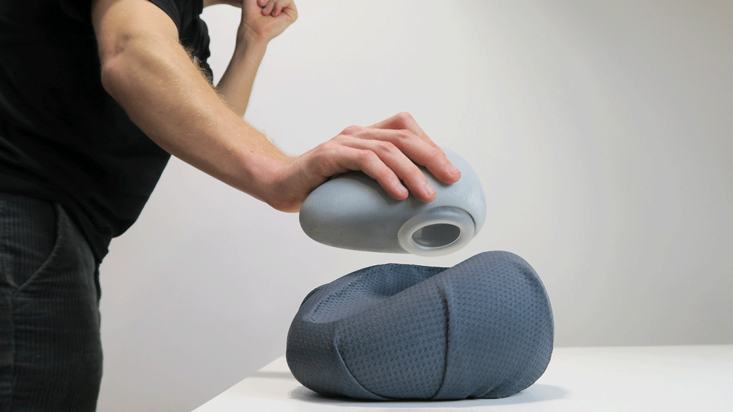 Alt text: A person using a uniquely shaped, gray, handheld device with a hollow opening to interact with a soft, dark gray cushion-like object on a table.