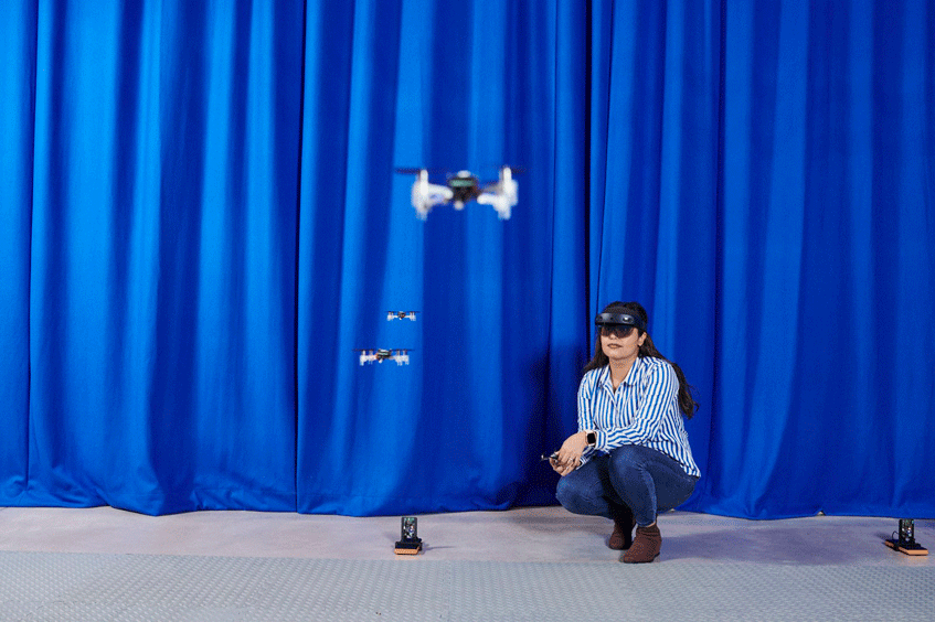 A researcher crouches by a flying drone robot in the robotics hangar