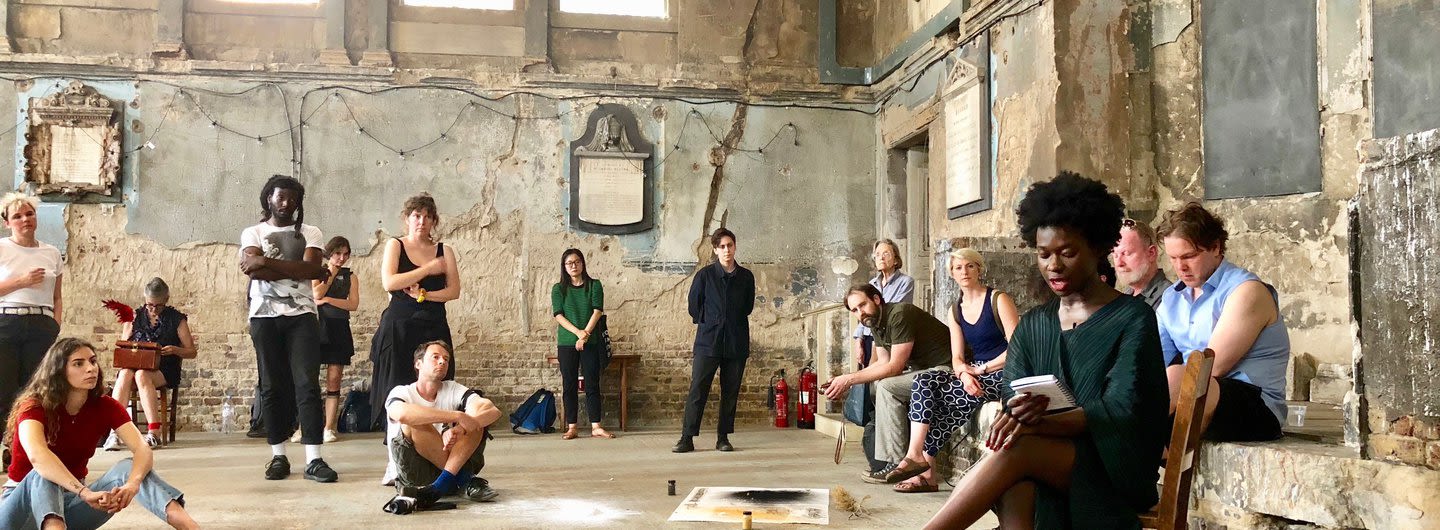 Research students grouped in a asylum, a dilapidated exhibition space, surround a person reading.