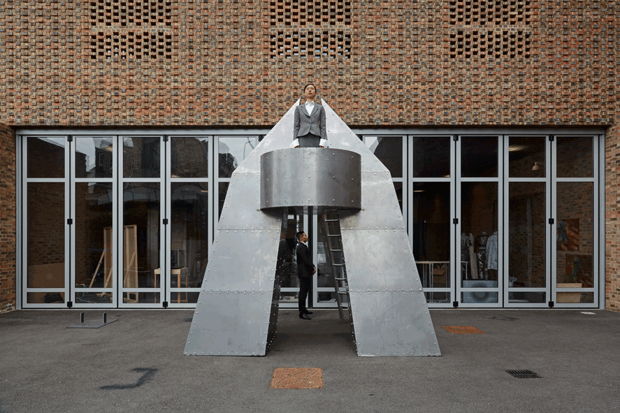 A large futuristic metal sculpture shaped like a rocket, with a person in a grey suit standing on its central platform