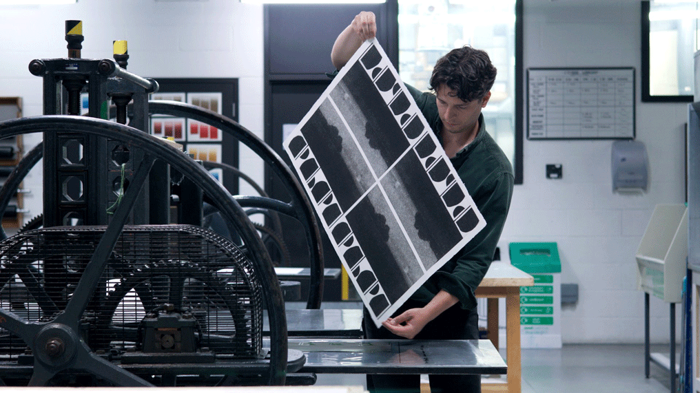 A student holds a large print next to a printing press